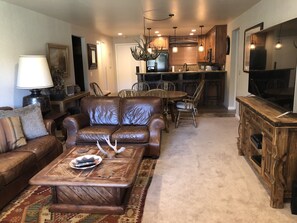 Sala de estar com sofá de couro marrom, mesa de centro e tapete. Há uma área de jantar com cadeiras e mesa de madeira. Uma cozinha é visível ao fundo, com armários de madeira e uma geladeira.
