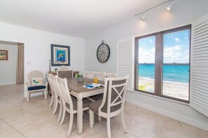 A dining area with a wooden table and white chairs, a clock on the wall, and a view of the beach through large windows. - Georgetown Villas #218 - 2BD Direct Oceanfront (Georgetown)