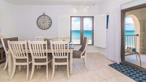 A dining area with a table and chairs, a wall clock, and a view of the sea through a window.