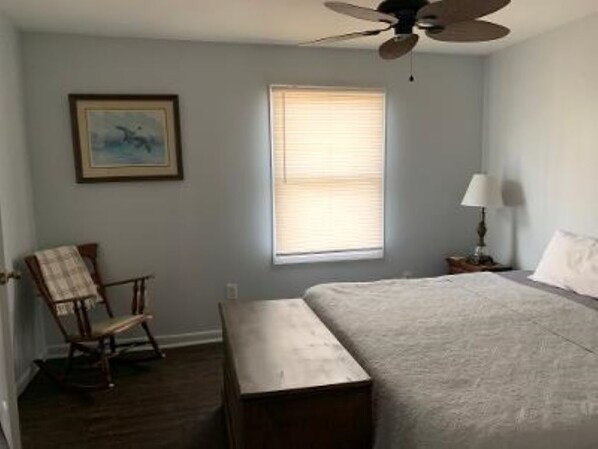 A bedroom with a bed, a chair, a small table, a ceiling fan, a window with blinds, and a wall-mounted lamp.