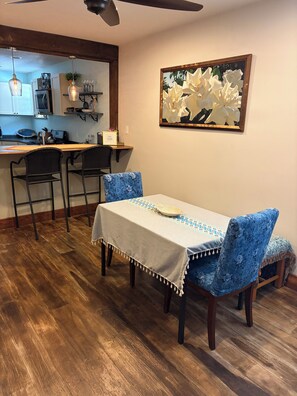 A dining area with a table set for four, flanked by blue upholstered chairs, a wooden floor, and a kitchen in the background.