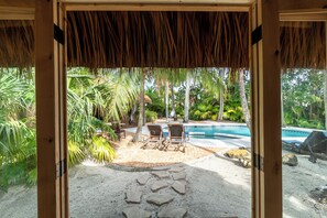 A view through a wooden-framed doorway into a tropical outdoor area with a pool, lounge chairs, and palm trees.
