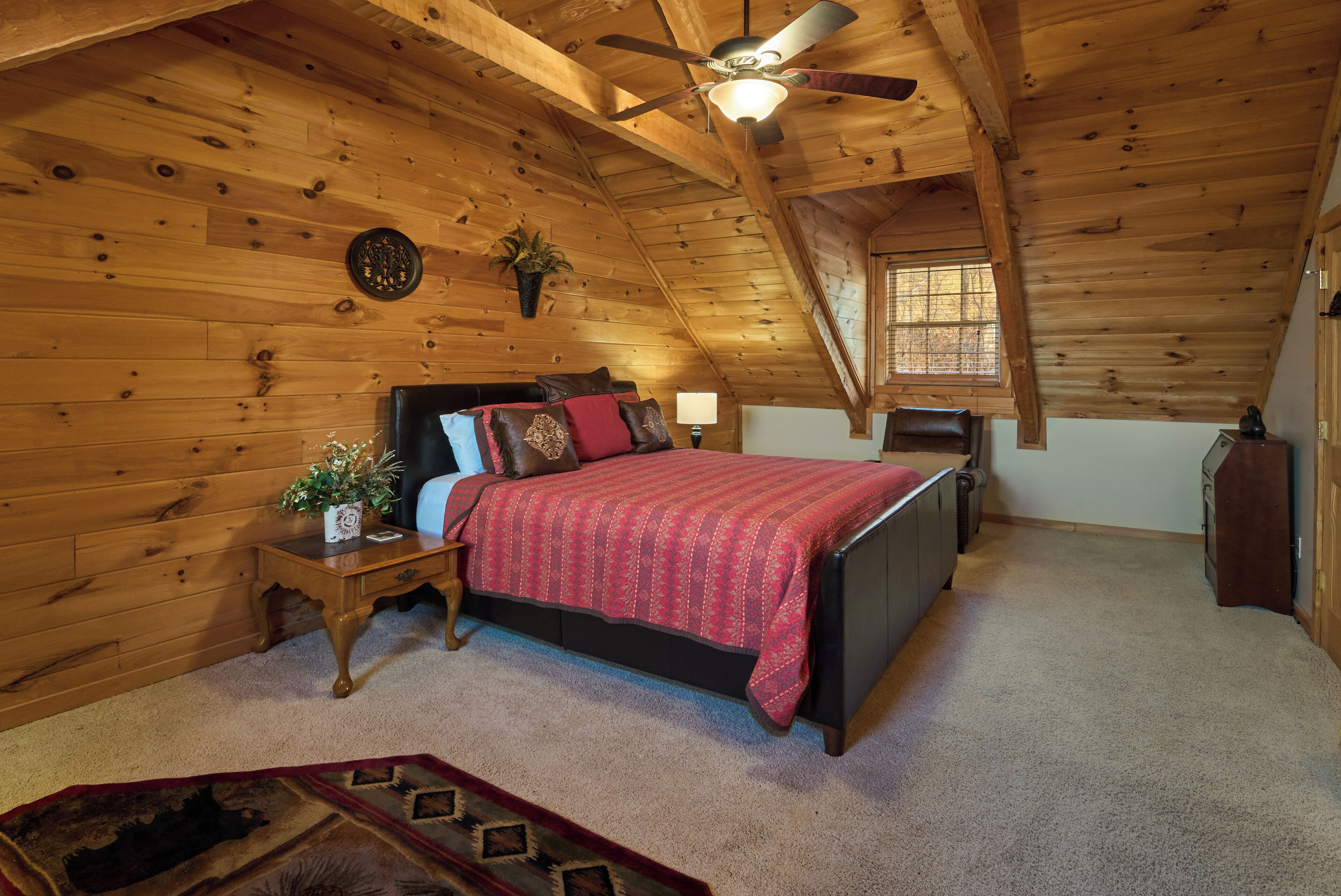 A bedroom with a wooden ceiling, a bed with a red blanket, a nightstand with a lamp, and a window with blinds.