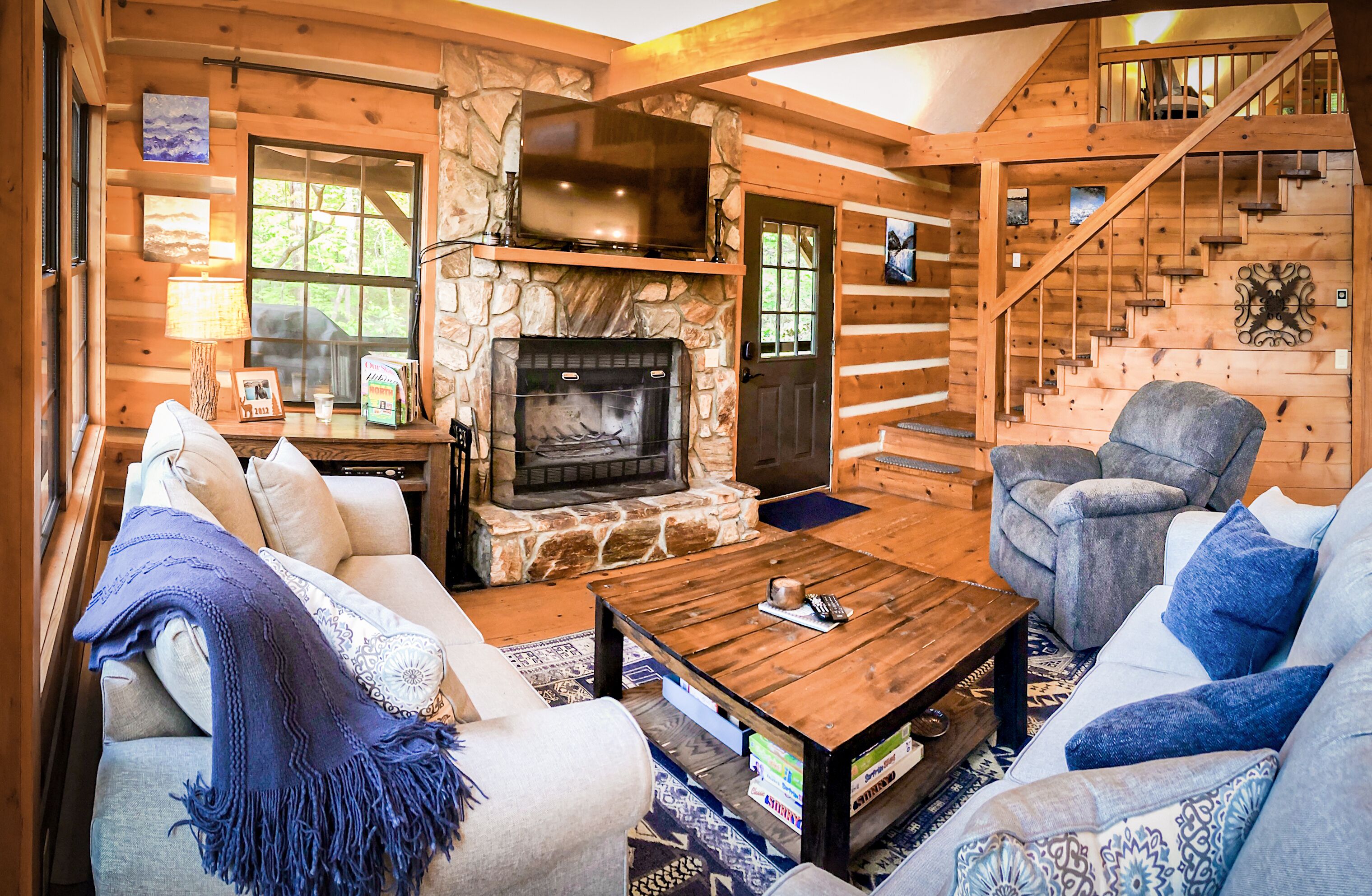 A log cabin interior with a stone fireplace, wooden walls, a sofa, a chair, and a staircase.