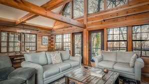 Ein Holzblockhaus-Innenraum mit zwei grauen Sofas, einem Holztisch und großen Fenstern mit Blick auf Bäume.