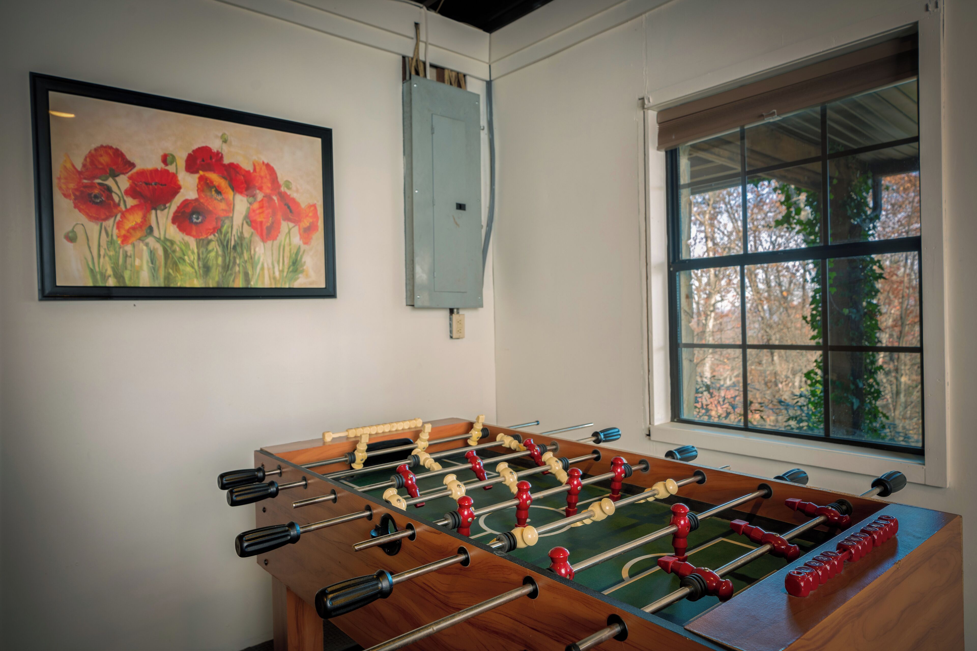 A room with a foosball table, a painting of red flowers, and a window with a view of trees.