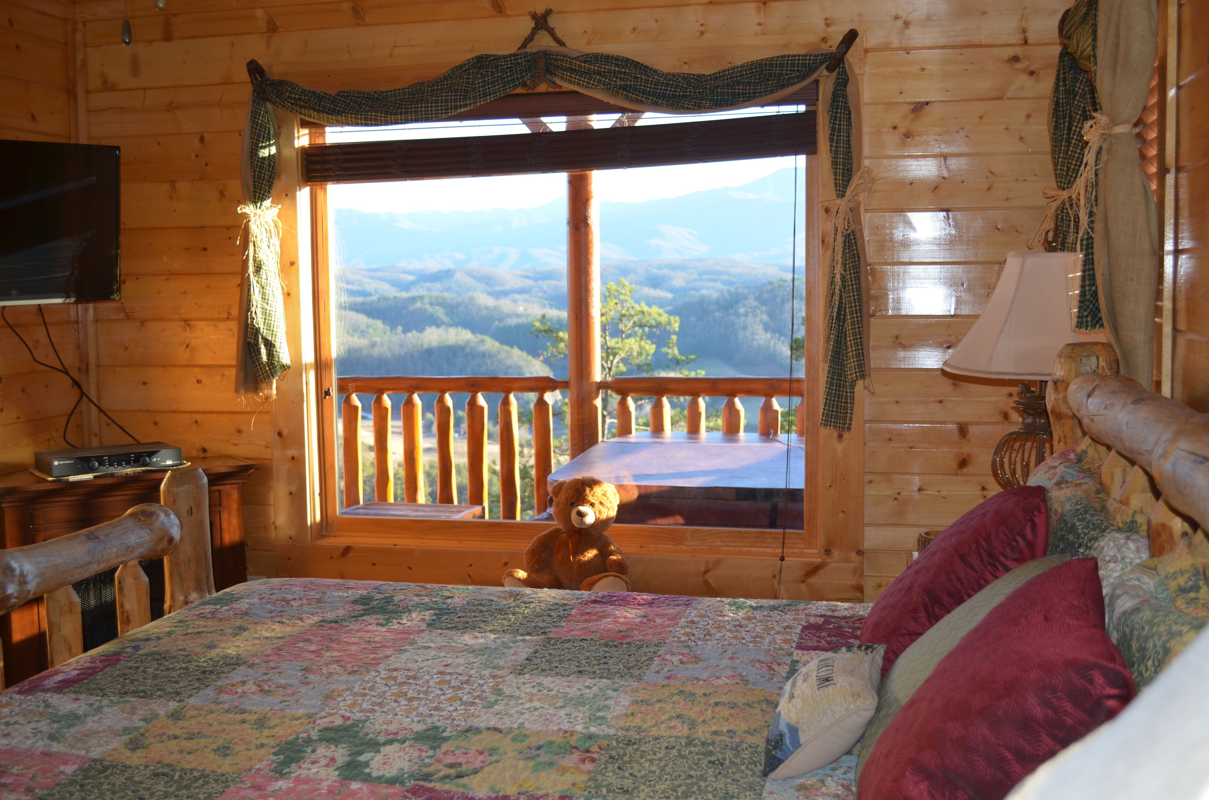 Ein Zimmer in einer Holzhütte mit großem Fenster, das einen Bergblick bietet, einem Bett mit floralem Quilt und einem Teddybären auf der Fensterbank.