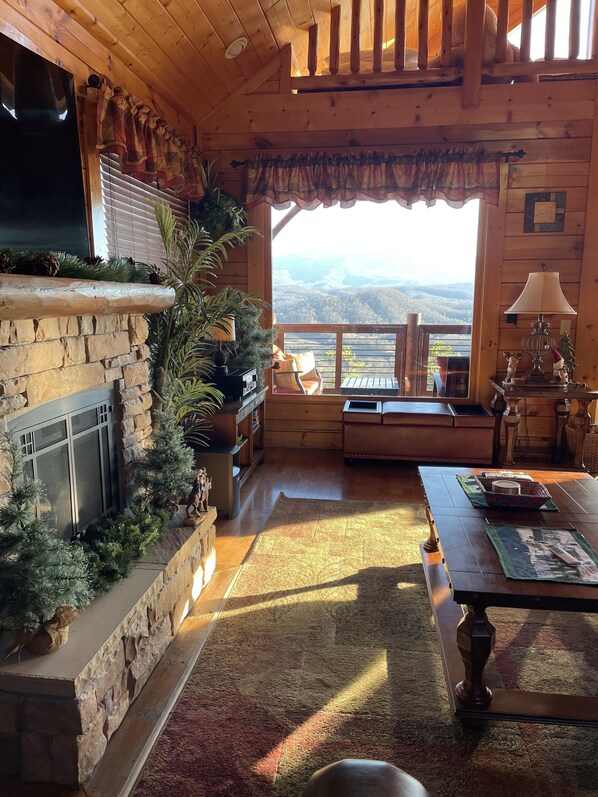 A cozy cabin interior with a stone fireplace, wooden ceiling, and a view of mountains.