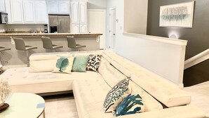 A modern living room with a beige sectional sofa, a small round table, and a kitchen area with white cabinets and a granite countertop. - BEAUTIFUL 3BR TOWNHOUSE w/ POOL and JACUZZI STEPS FROM BEACH - BOOK NOW! (South Padre Island)