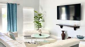 A modern living room with a sectional sofa, a glass coffee table, a potted plant, and a flat-screen TV mounted on the wall. - BEAUTIFUL 3BR TOWNHOUSE w/ POOL and JACUZZI STEPS FROM BEACH - BOOK NOW! (South Padre Island)