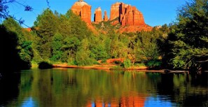 A natural landscape with a calm lake reflecting red rock formations and dense green foliage.