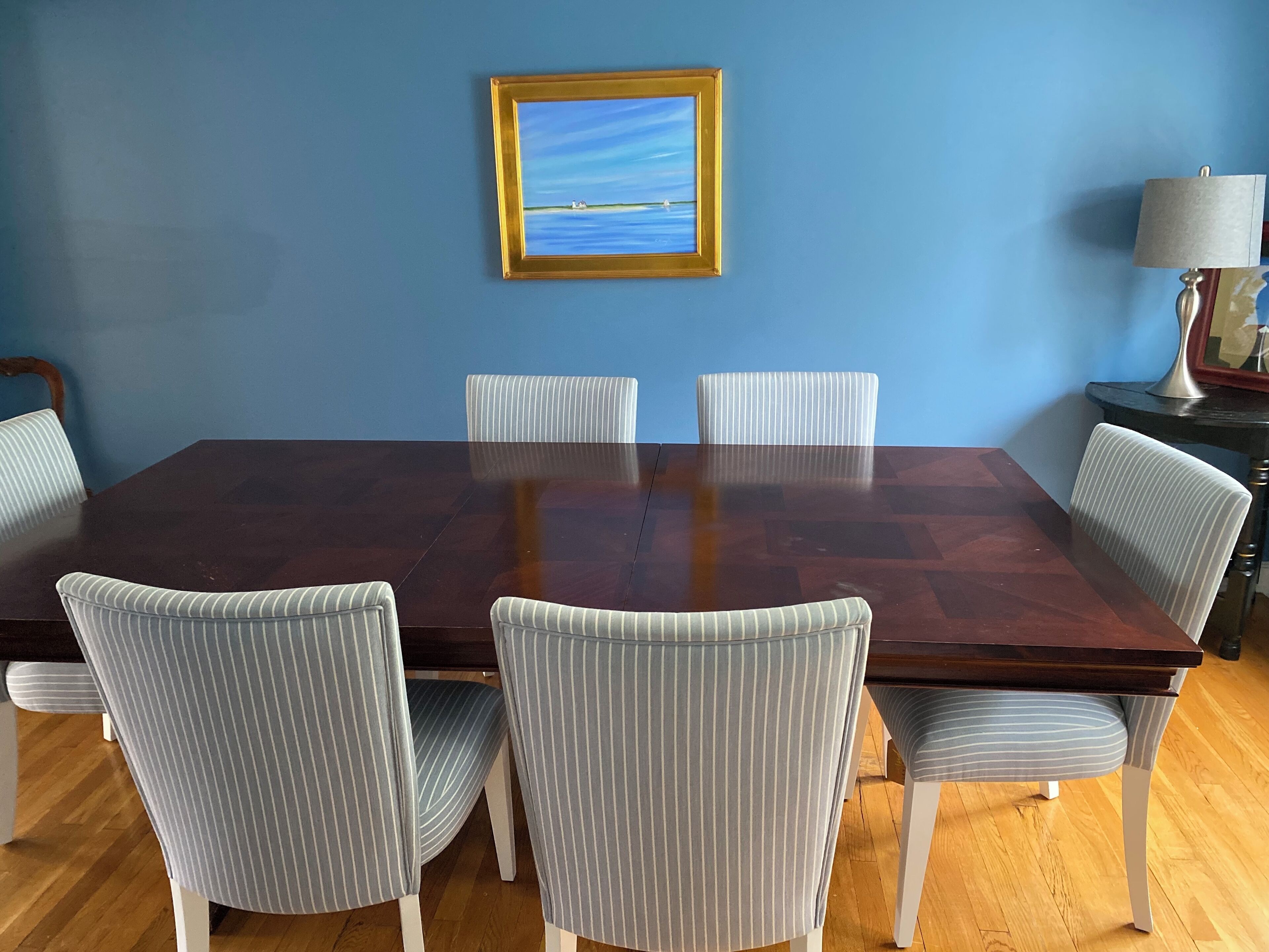 A dining room with a large wooden table surrounded by six striped chairs, a framed picture on the wall, and a lamp on a side table.