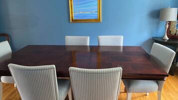 A dining room with a large wooden table surrounded by six striped chairs, a framed picture on the wall, and a lamp on a side table.