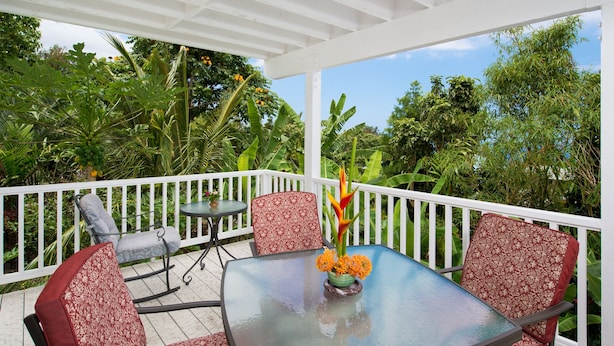A covered outdoor patio with a glass table and red patterned chairs, surrounded by lush greenery.