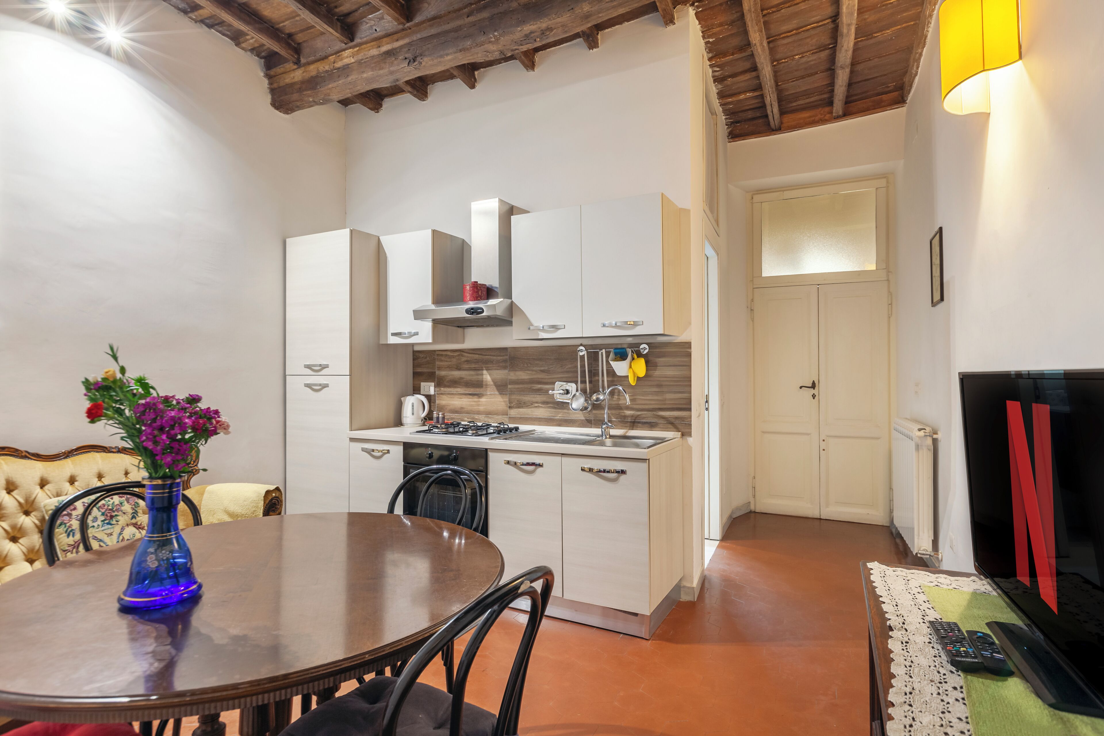 A compact kitchen with white cabinets, a stove, and a sink. There is a dining table with chairs and a vase with flowers.