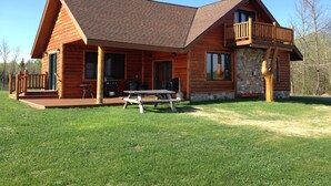 A log cabin with a stone foundation, a balcony, and a picnic table in the front yard.