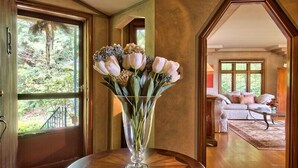 A room with a wooden floor, a round table with a vase of flowers, and a view of a garden through a window. - On The Beach! Elegant Waterfront Manor 'The Pearl on Oyster Bay (Bremerton)