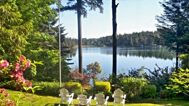 A serene lakeside garden with three Adirondack chairs facing a calm lake, surrounded by lush greenery and tall trees. - On The Beach! Elegant Waterfront Manor 'The Pearl on Oyster Bay (Bremerton)