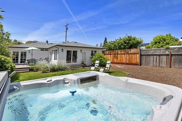 A backyard with a hot tub, a patio with chairs, a wooden fence, and a house.