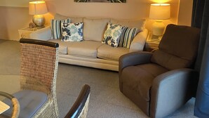 Ein Wohnzimmer mit einem beigen Sofa, zwei gemusterten Kissen, einem Korbstuhl und einem braunen Sessel. An der Wand hängt ein Gemälde einer Strandansicht, und auf beiden Seiten des Sofas stehen Lampen.