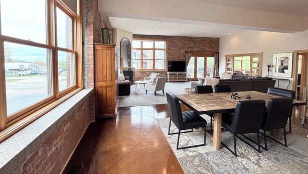 A spacious living area with a large wooden dining table, comfortable seating, a fireplace, and large windows offering a view of the outdoors.