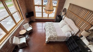 A spacious bedroom with a large bed, a sitting area with a chair and a small table, a hanging light fixture, and a view of the street through large windows.