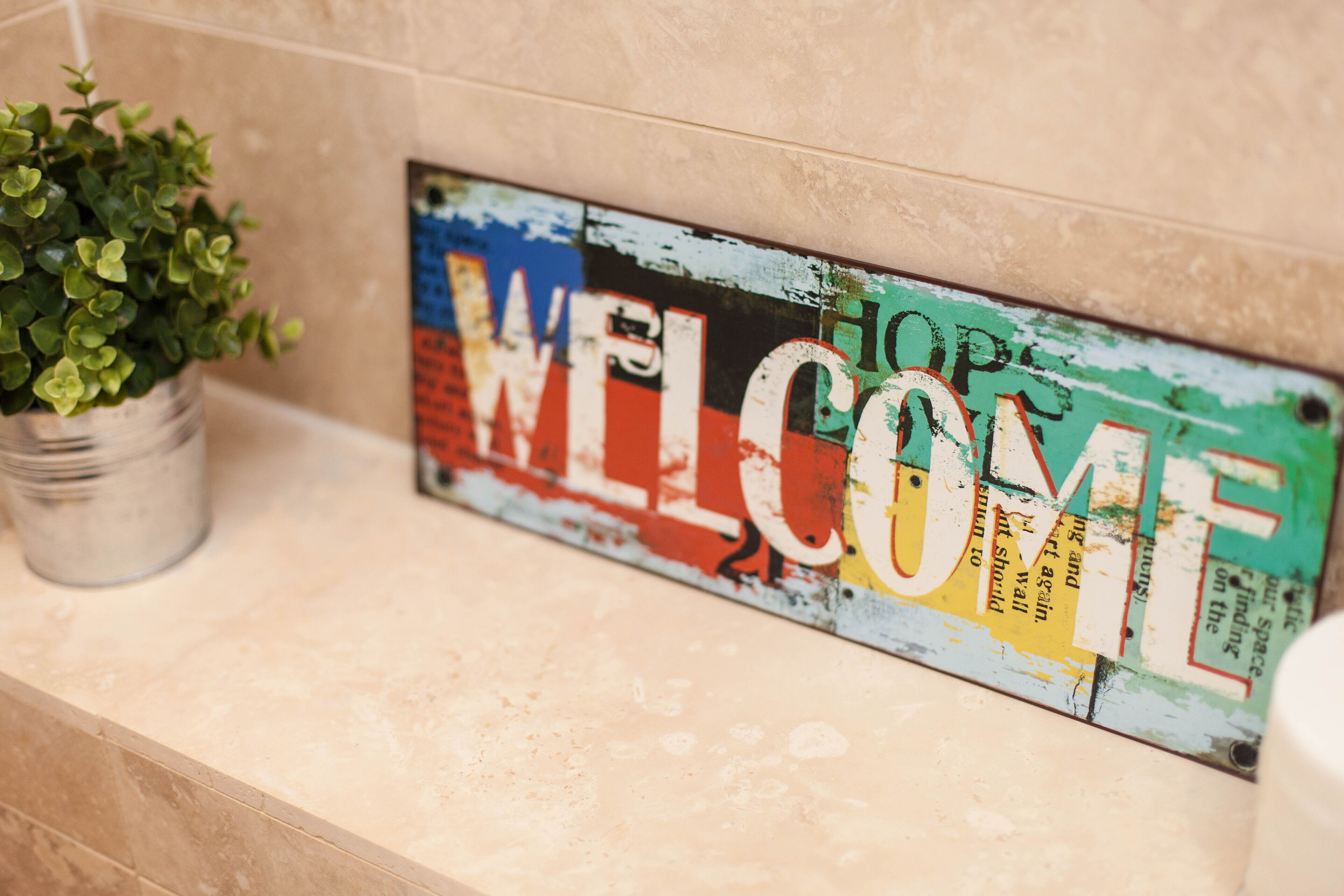 Un letrero de bienvenida con la palabra 'WELCOME' en letras grandes, una pequeña planta en maceta y una pared con azulejos.