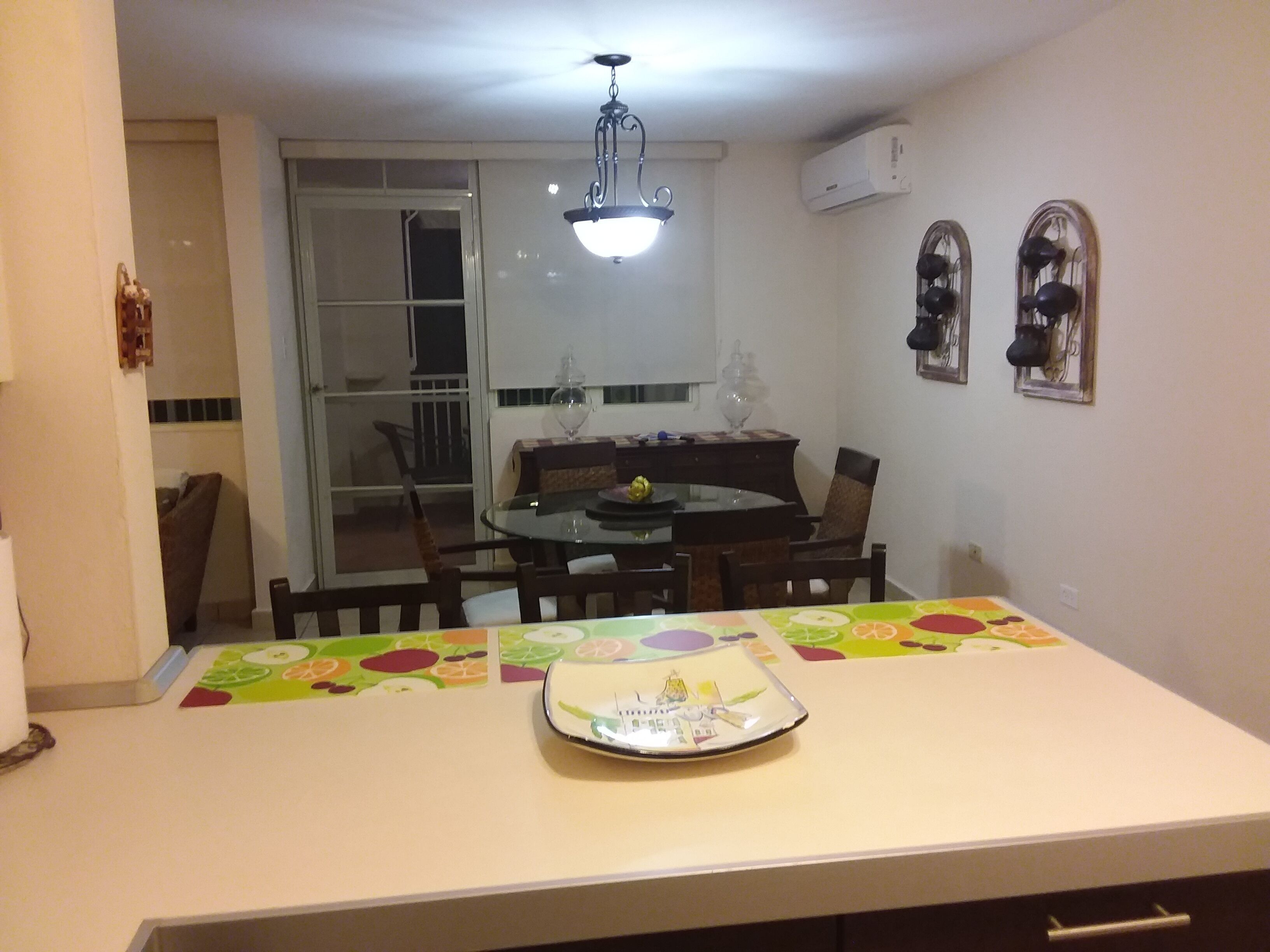 A dining area with a glass table, chairs, and a colorful tablecloth. There is a ceiling light fixture above the table.