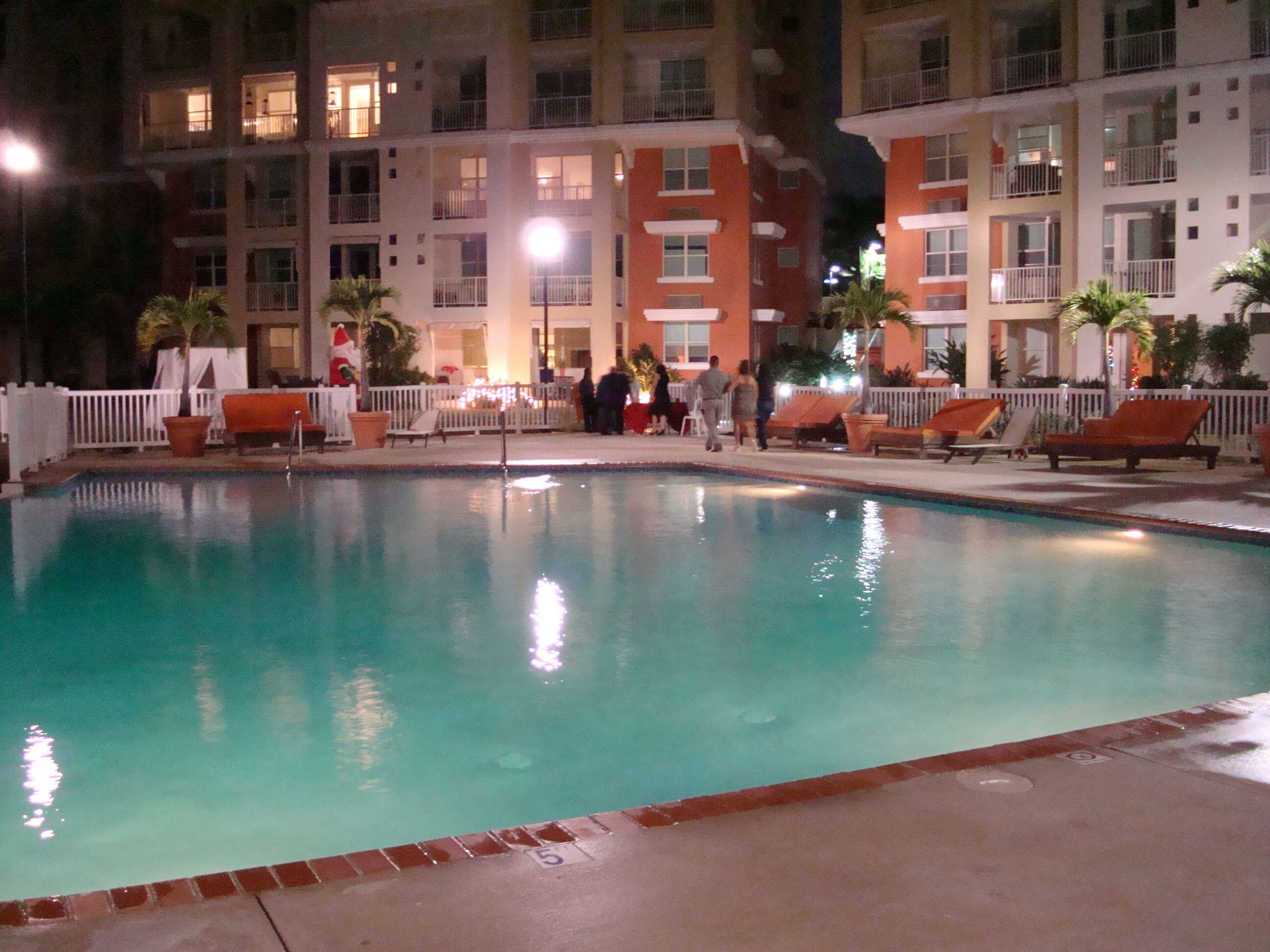 Une zone de piscine éclairée la nuit, avec des chaises longues et un bâtiment en arrière-plan.