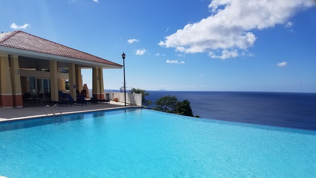 Una piscina azul cristalina con un indicador de profundidad de 3 pies, un área de estar cubierta con sillas y mesas, y vista al océano.