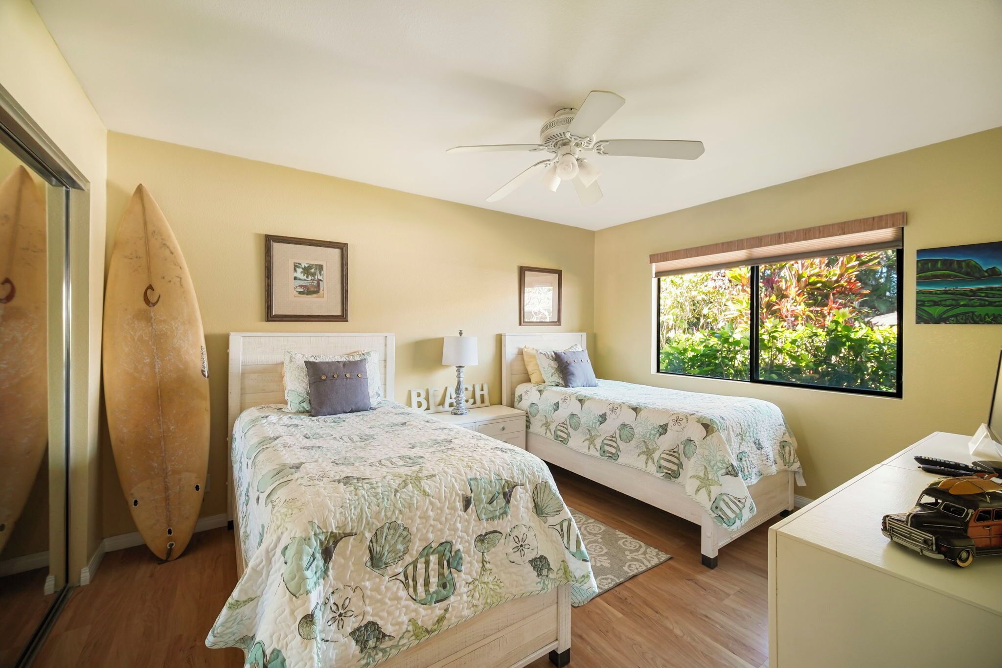A bedroom with two beds, a ceiling fan, a surfboard, and a window with a view of greenery.