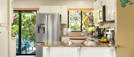 Una cocina moderna con armarios blancos, un refrigerador de acero inoxidable, una encimera de granito y vistas a un jardín a través de una puerta corrediza de vidrio.