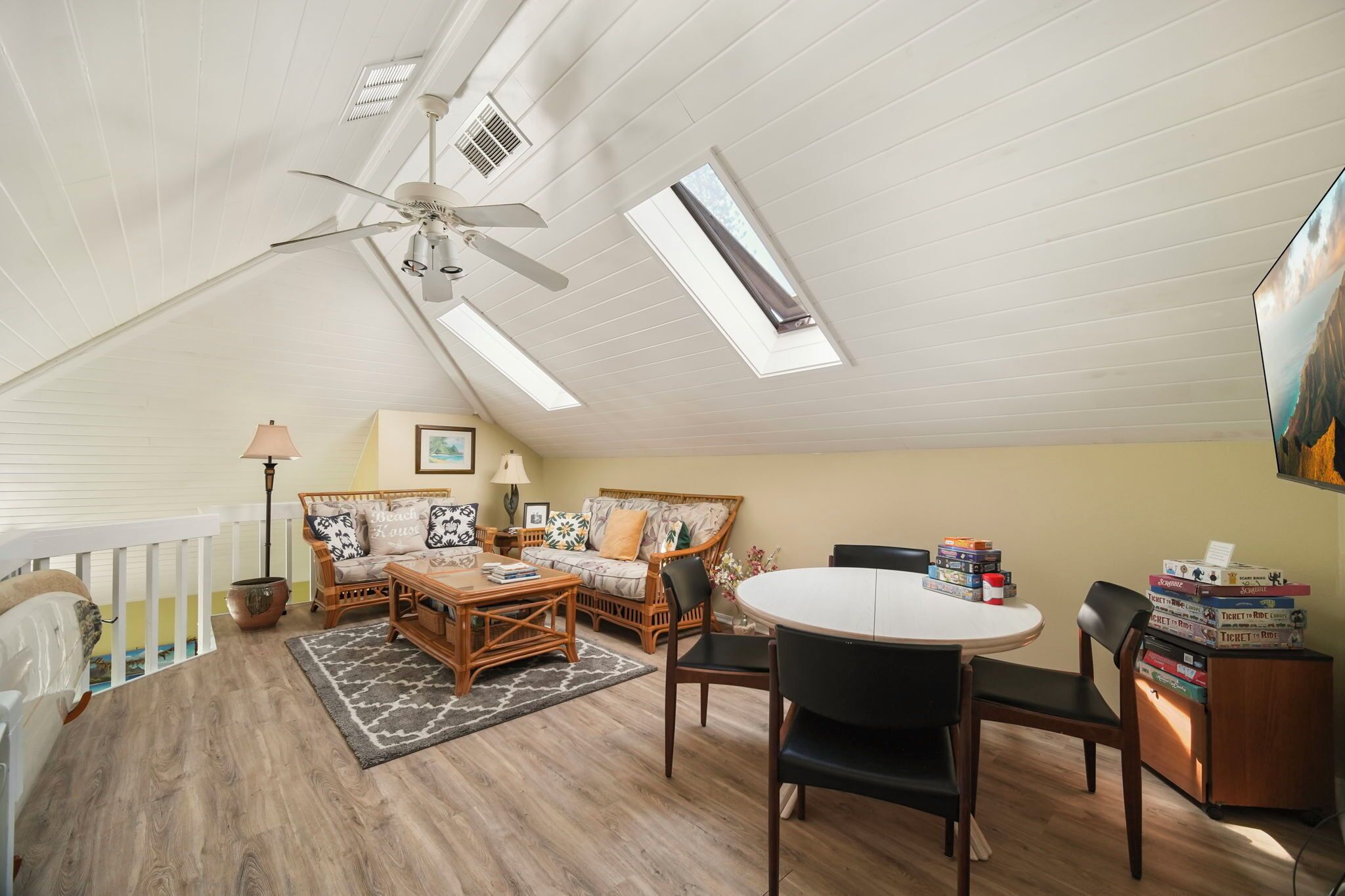 A spacious attic room with a high ceiling, skylights, a ceiling fan, a dining table with chairs, a sofa, and a bookshelf.