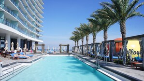 Un espace piscine doté de chaises longues et de palmiers, situé à côté d’un immeuble à plusieurs étages avec des balcons.