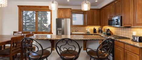 A kitchen with wooden cabinets, a stainless steel refrigerator, a microwave, and a granite countertop. There are three bar stools and a dining table with chairs.