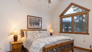 Ein Schlafzimmer mit einem Holzbett, zwei Nachttischen, einem gerahmten Bild an der Wand und einem großen Fenster mit Blick auf Bäume und Berge.