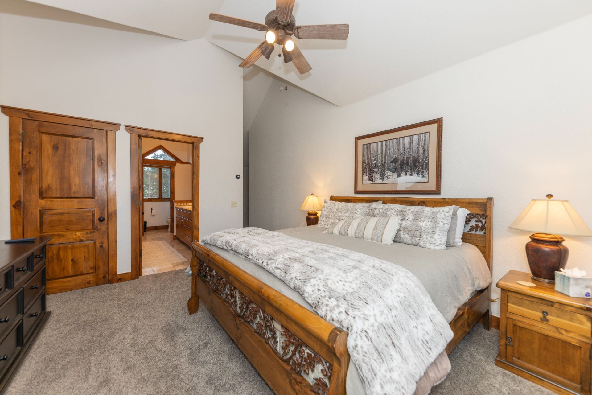 A bedroom with a wooden bed, a ceiling fan, a framed picture on the wall, and a nightstand with a lamp.