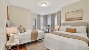 A hotel room with two beds, a desk, a chair, a lamp, a painting, and a window with curtains.