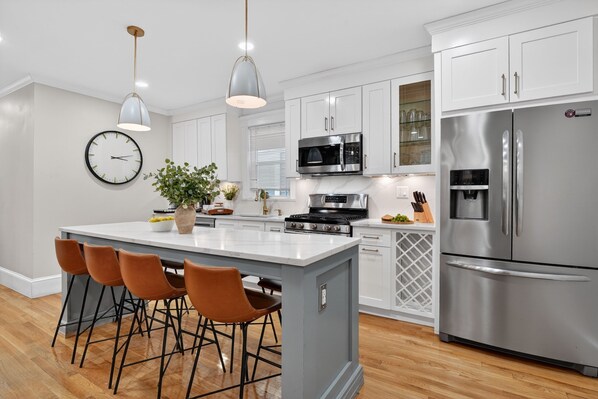 A modern kitchen with white cabinets, stainless steel appliances, a central island with bar stools, and pendant lighting.