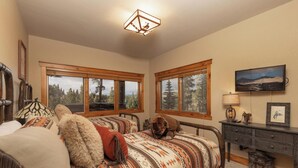 A bedroom with two beds, a wooden dresser, a mounted TV, and large windows with a forest view. - Slopeside Saddlewood: True Ski-in/Out, Private Hot Tub, Grill, Shuttle (Breckenridge)