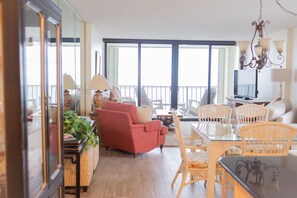 A spacious living room with a red sofa, a glass dining table, and wicker chairs. There is a TV and a chandelier.
