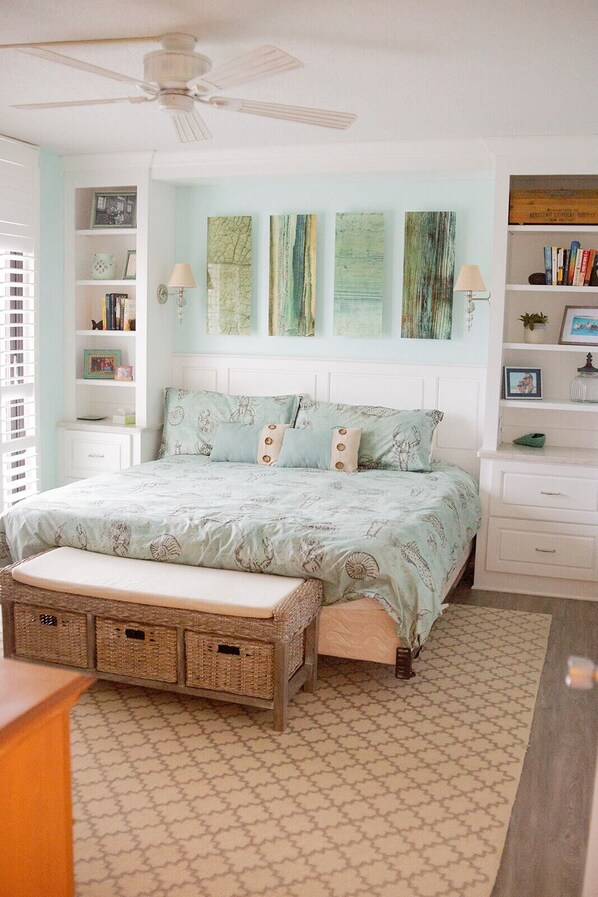 A bedroom with a bed, a bench, a ceiling fan, wall art, and a bookshelf.