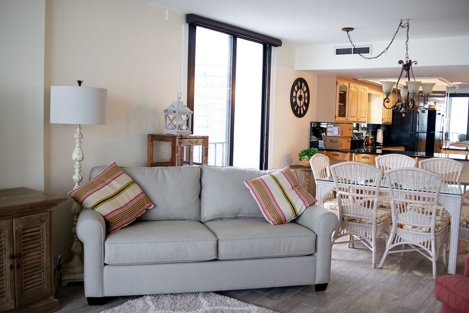A living room with a sofa, a dining area with wicker chairs, a kitchen in the background, and a large window.