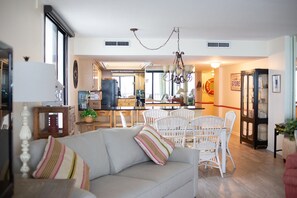 A spacious living area with a sofa, dining table, and chairs. A kitchen with a refrigerator and stove is visible in the background.
