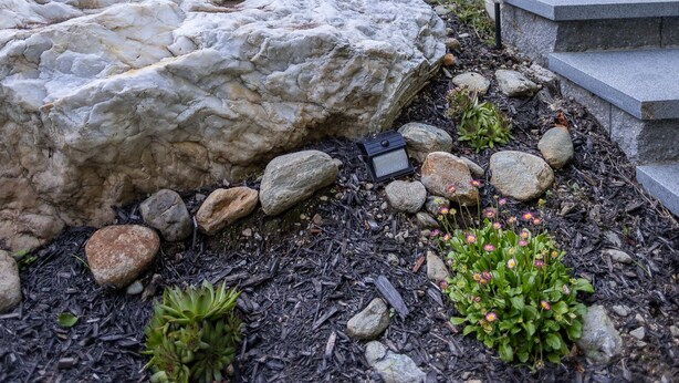 Una casa moderna con fachada de piedra, un balcón con flores y un jardín bien cuidado con rocas y plantas.