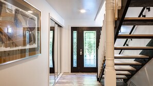 Un pasillo con piso de madera, una puerta negra con paneles de vidrio y una escalera con barandilla blanca.