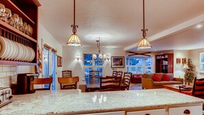A spacious kitchen with a large island, wooden chairs, and a dining table. There are hanging light fixtures and a view of the city through large windows.
