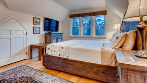 A bedroom with a wooden bed, a desk, a chair, a TV, and a window with a mountain view.
