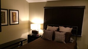 Un dormitorio con una cama, una mesita de noche con lámpara, un banco y dos cuadros en la pared.