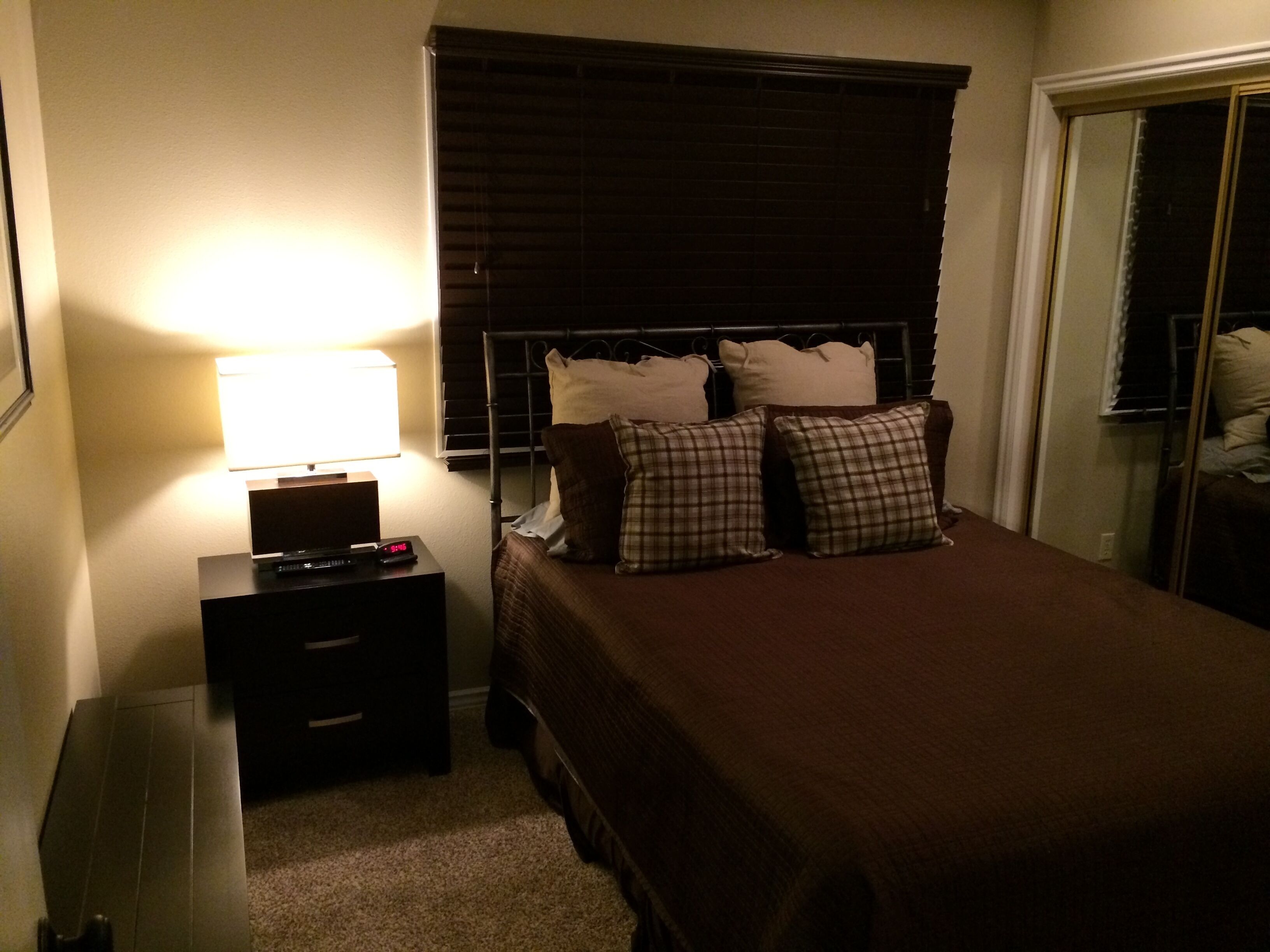 A bedroom with a bed, a nightstand with a lamp, a window with blinds, and a door.